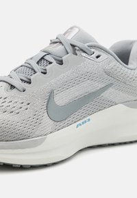Šedé športové topánky s priedušným vrchom zo sieťoviny, štruktúrovanými prekrytím a bielou podošvou. Obsahuje výrazný šedý „swoosh“ a modré logo „AIR“. 