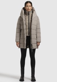 Manteau matelassé de couleur beige clair, doté d'un col haut, d'une fermeture éclair et de poches latérales. Porté par-dessus une couche intérieure ajustée et un legging noir.