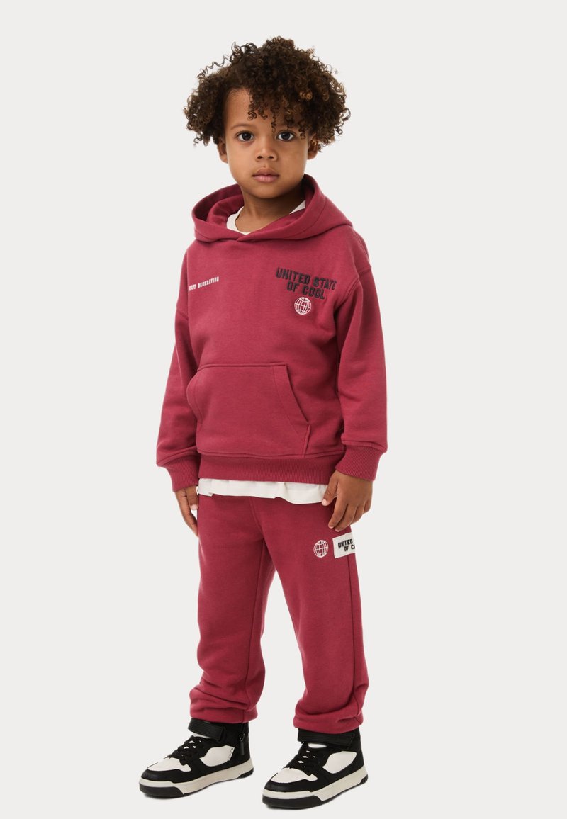 Bordeaux hoodie en sweatpants set, katoenmateriaal, voorzak, ribgebreide manchetten, met grafische tekstaccenten, gecombineerd met zwart-witte sneakers.