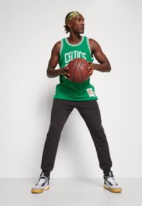 Grünes Basketballtrikot mit dem Text "CELTICS", schwarze Jogginghose und graue Turnschuhe mit gelben Akzenten, hält einen Basketball, dynamische Pose.