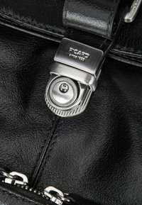 Detalle de bolsa de cuero negro con un cierre metálico plateado con el logotipo grabado "PCARD" y un intrincado diseño circular. Acentos de cremallera visibles en la parte inferior.