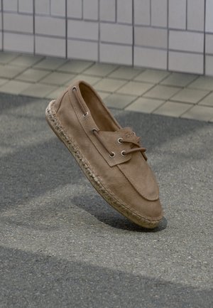 Lysebrun ruskind loafer med syning i sålen og snøredetalje, vist svævende over tekstureret asfalt og flisebelagt baggrund.