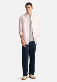 Helles rosa Button-Up-Hemd über einem grauen T-Shirt, kombiniert mit marineblauen Hosen und beigen Turnschuhen. Das Hemd hat eine Brusttasche und hochgekrempelte Ärmel.