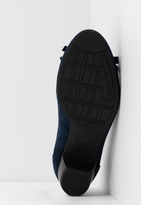 Semelle d'une chaussure habillée bleu marine avec motif de bande de roulement et petit talon, placée verticalement contre un fond blanc en angle.