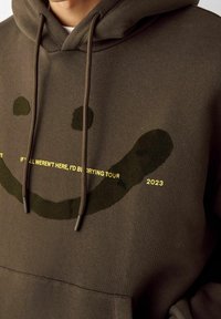 Felpa marrone con cappuccio con una grande grafica di una faccina sorridente in verde scuro, con testo giallo che recita "IF I ALL WEREN'T HERE, I'D BE CRYING TOUR 2023."