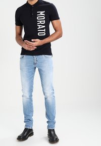 Svart polo-tröja med vertikal vit text "MORATO", tillsammans med ljusblå jeans och svarta ankelstövlar. Enkel design, figursydd form.