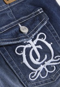 Τζιν τσέπη με ασημένιο κουμπί, διακοσμημένη με λευκά κεντημένα αρχικά και μικρές πολύτιμες πέτρες, σε σκουρό μπλε ύφασμα με αντίθετες ραφές.