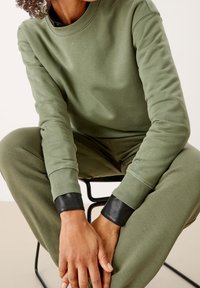 Maglione verde in cotone con maniche lunghe, dotato di polsini in pelle nera. Il design è minimale con scollo rotondo e una texture morbida.