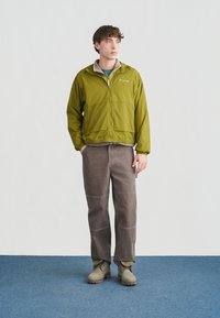 Junger Mann steht auf einem blauen Teppich, trägt eine grüne Jacke zum Reißverschluss, beige Stiefel und braune Hosen, schaut seitlich vor einem schlichten weißen Hintergrund.