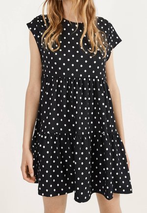 Kobieta w czarnej sukience z krótkim rękawem w białe groszki, z falbanami i luźno opadającym dołem.