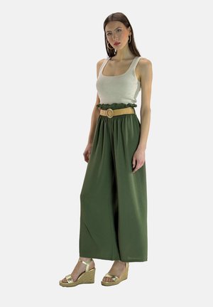 Grüne weit geschnittene Hose mit rüschigem Bund und einem geflochtenen Strohhut, kombiniert mit einem weißen Tanktop und metallic Wedge-Sandalen.
