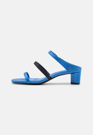 Blue and black heeled sandals featuring three straps, smooth texture, square toe, and a block heel for stability. Subtle branding detail included.