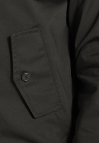 Ciemnozielona tkanina o gładkiej fakturze, z kieszenią z klapką zabezpieczoną okrągłym, czarnym guzikiem, obszytymi krawędziami i subtelnym połyskiem.