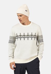 Hellgrauer Pullover mit geripptem Rundhalsausschnitt und schwarzem geometrischem Muster auf der Brust. Kombiniert mit einer passenden gerippten weißen Mütze.