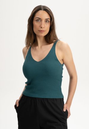 Frau mit langen braunen Haaren, die ein türkisfarbenes, geripptes, ärmelloses Oberteil und schwarze Hose trägt, steht mit den Händen in den Taschen vor einem schlichten Hintergrund.