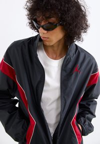 Black and red windbreaker jacket with white detailing, featuring a zip front and a logo on the upper left. Model wears sunglasses.