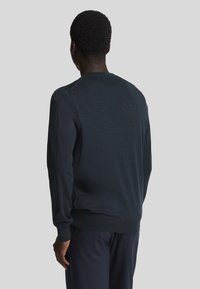 Maglione blu scuro con maniche lunghe e collo rotondo. Tessuto morbido, polsini e orlo ribattuti, e dettagli di cucitura minimi. Indossato di spalle.