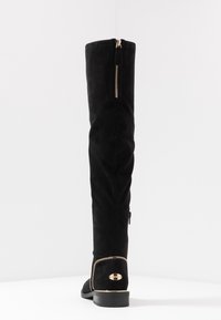 Sorte ruskind over knæstøvler med en gulddetalje med lynlås bagpå og en metallisk accent på hælen. Flad sål og rund tå-design.