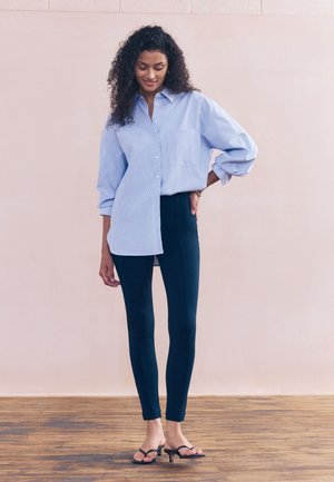 Frau mit lockigem Haar, die ein lockeres hellblau gestreiftes Hemd, dunkle, eng anliegende Hose und schwarze Sandaletten mit Absatz trägt, steht auf einem Holzboden.