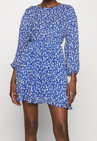 Kvinna i en blå blommig långärmad minikjol med elastisk midja och rund halsringning mot en enfärgad bakgrund.