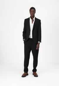 Black suit jacket with notch lapels, white shirt, relaxed-fit black trousers, and brown leather shoes, on a model against a white background.