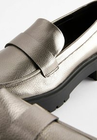 Mocasines metálicos plateados con un acabado texturizado, que presentan una correa ancha en el empeine y una suela negra resistente. Detalle de diseño geométrico visible.