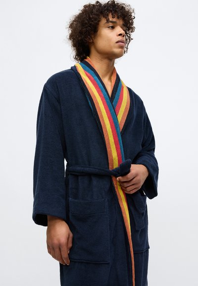 Mees, kes kannab mereväe sinist terri-froteest hommikumantlit, millel on mitmevärviline triibuline krae, hoides rihma, seistes ühetaolise tausta ees.