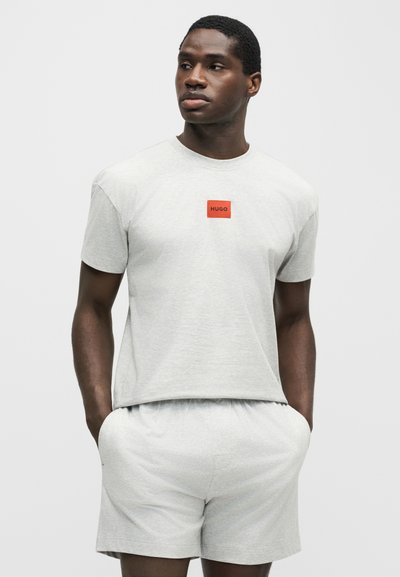 Homme portant un t-shirt gris clair à manches courtes avec un logo orange et un short assorti, mains dans les poches, regardant légèrement sur le côté.