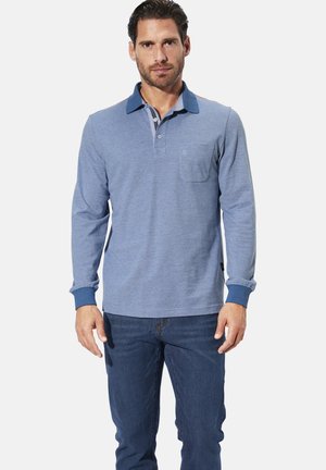 Langärmliges blaues Poloshirt mit einem kontrastierenden dunkelblauen Kragen, kleiner Brusttasche und taillierten Manschetten. Getragen mit dunklen Jeans.