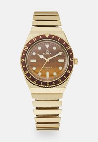 Montre à quartz Timex avec un boîtier et un bracelet en métal doré. Le cadran marron affiche des index horaires blancs et une lunette bordeaux avec des repères de minutes.