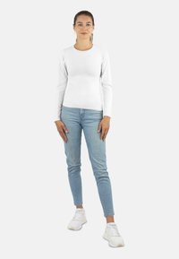 Frau steht frontal und trägt ein weißes langärmliges Shirt, hellblaue Jeans, weiße Sneakers und goldene Ohrringe vor einem schlichten Hintergrund.
