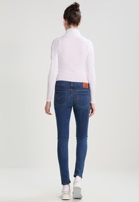 Jeans de mezclilla azul ajustados con bolsillos traseros y una etiqueta de marca de cuero marrón; combinados con un suéter de cuello alto blanco ceñido y zapatillas blancas.