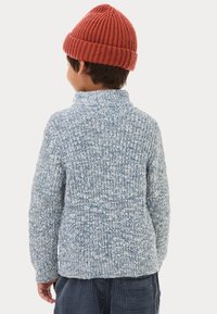 Maglione lavorato a maglia di colore azzurro chiaro con colletto alto, caratterizzato da un motivo testurizzato. Abbinato a un berretto a coste rosso. Indossato con pantaloni a strisce scure.