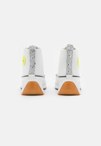 Witte hoge sneakers met een glinsterende zilveren hieltab, grijze zool en een ronde gele badge aan de zijkant. Gestructureerde stof, rubberen accenten.