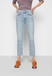 Ljust blå jeans med fransad nederkant, hög midja och fem fickor; bärs med orange remsandaler som framhäver kontrasten i färg och textur.