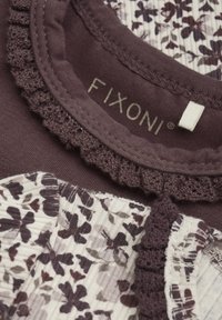 Lila bomullstopp med volangkrage, som har blommönster i nyanser av plommon och kräm. Märkesetikett synlig, textilstruktur längs kanterna.