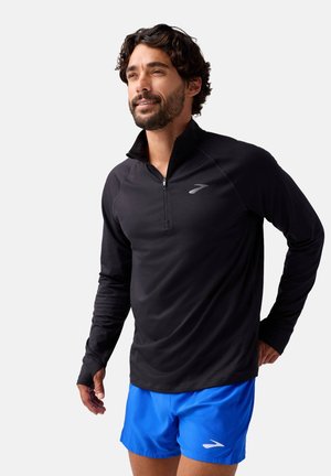 Black long-sleeve athletic top with a quarter zipper, featuring a logo on the chest. Worn with blue shorts. Smooth fabric texture.