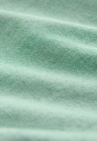 Tuvu skatā redzama zaļas austas auduma tekstūra ar mazām, vienmērīgi izvietotām diegām un smalkām krokām, kas rada maigas ēnas.