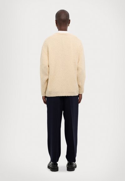 Beige gestrickter Pullover mit lockerer Passform, geripptem Saum und Bündchen, kombiniert mit dunkelblauen weit geschnittenen Hosen und schwarzen Schuhen. Ansicht von hinten.
