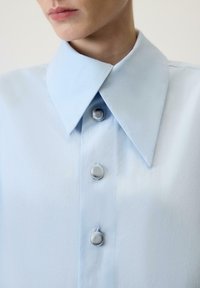 Gaiši zila krekla ar apkakli ar gludu tekstūru, lieli pelēki pogas un smailu apkakles dizains. Tuvplāna skats.