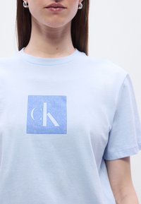 Ανοικτό μπλε βαμβακερό μπλουζάκι με τετράγωνο λογότυπο που απεικονίζει το στιλιζαρισμένο "cK" σε μπλε χρώμα. Κοντά μανίκια και λαιμόκοψη τύπου crew. Λείο ύφασμα.
