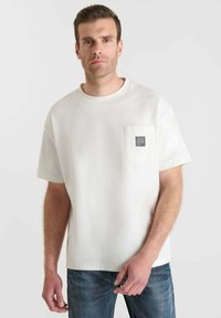 Uomo che indossa una maglietta bianca a maniche corte con taschino e jeans blu, in piedi contro uno sfondo bianco.