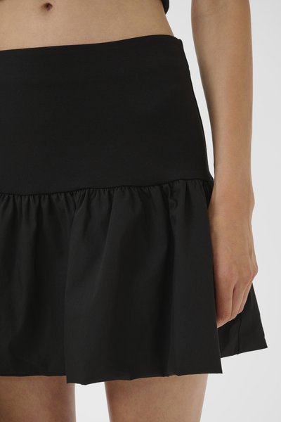 Femme portant une jupe courte noire avec une taille cintrée et une partie inférieure froncée et évasée, debout devant un fond uni.