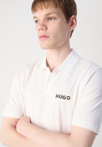 Hvit polo skjorte med klassisk krage, med en to-knapps placket og svart brodert "HUGO" logo på venstre bryst.