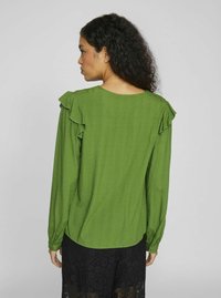 Green long-sleeve blouse with ruffle accents at shoulders, textured fabric, rounded neckline, and a slightly curved hem.