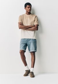 Béžové dvojfarebné tričko s nápisom „BROOKLYN“, v kombinácii s modrými džínsovými šortkami. Model má na sebe biele ponožky a hnedé semišové tenisky.