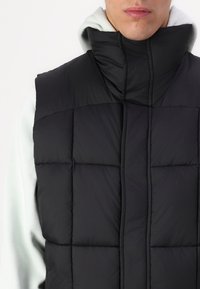Black puffer vest with high collar, featuring a quilted design and glossy finish, layered over a light gray hoodie.