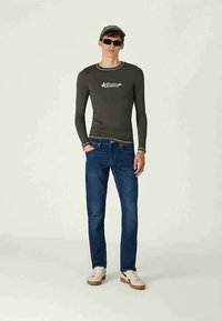 Oliven grønn langermet skjorte med hvitt logo, blå jeans, hvite sneakers med beige detaljer, og solbriller, står mot en lys bakgrunn.