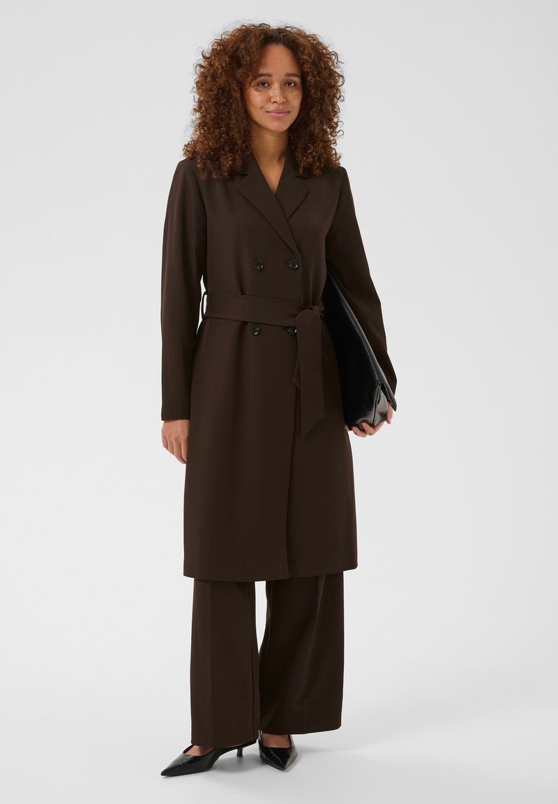Femme aux cheveux bouclés portant un manteau marron foncé ceinturé, un pantalon assorti et des escarpins noirs, tenant une grande pochette noire.