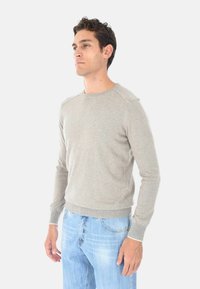 Uomo in maglione a maniche lunghe grigio chiaro e jeans azzurri chiari in piedi contro uno sfondo bianco uniforme, che guarda di lato.